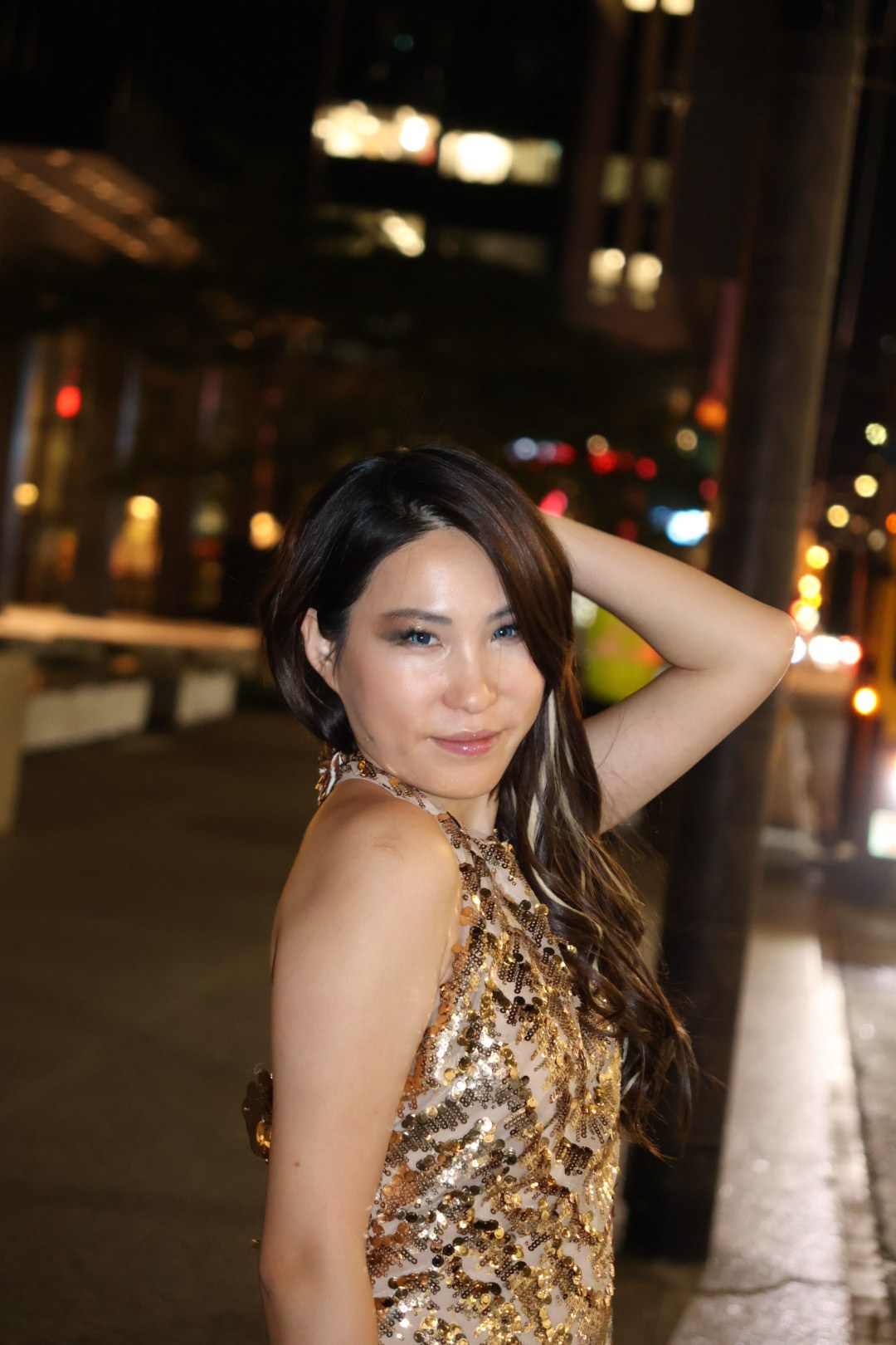 Enyu in a gold sequinned dress with one arm raised against warm evening lights