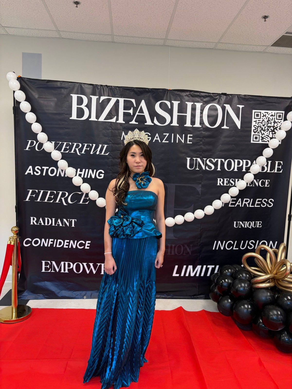 Enyu in a blue gown and crown at a BIZFASHION Magazine event backdrop